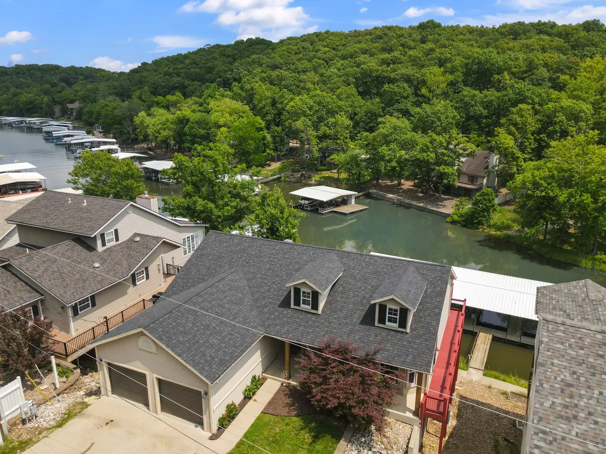 This exterior image captures the house with a sloped roof nestled among lush greenery, set beside the calm water of the river. In the foreground, nearby boat docks are visible, reinforcing the lakefront setting and proximity to water activities.