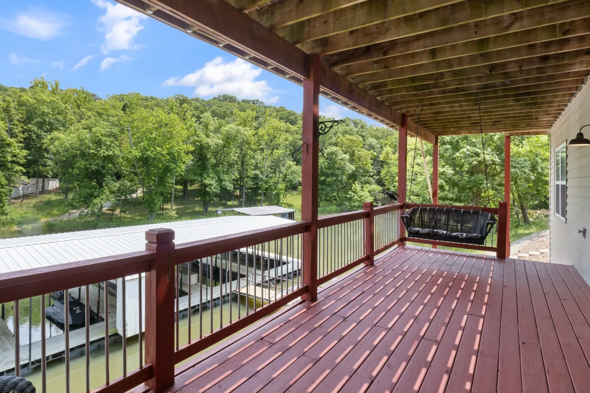 This exterior view centers on a swinging bench overlooking the waterfront from a wooden deck, framed by lush green trees. The scene captures a peaceful moment of relaxation and offers a glimpse of the natural lakeside environment and dock area.