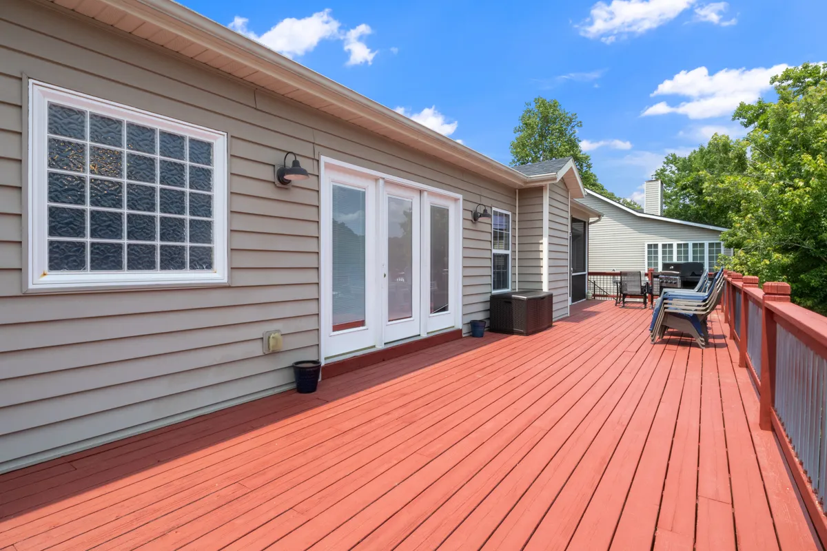 A broad view of the wooden lakeside deck, with sliding glass doors leading from the interior, surrounded by green trees and neighboring houses visible in the background. The deck is furnished for gatherings and suggests ample space for outdoor entertaining.