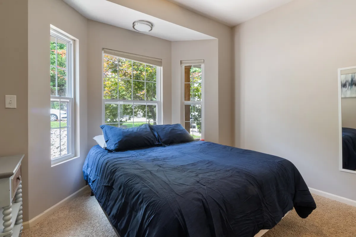 A serene bedroom with blue bedding on a plush bed, large windows letting in natural light, and a mirror on the wall. The ambiance is cozy and peaceful, perfect for restful nights or quiet afternoons spent reading.
