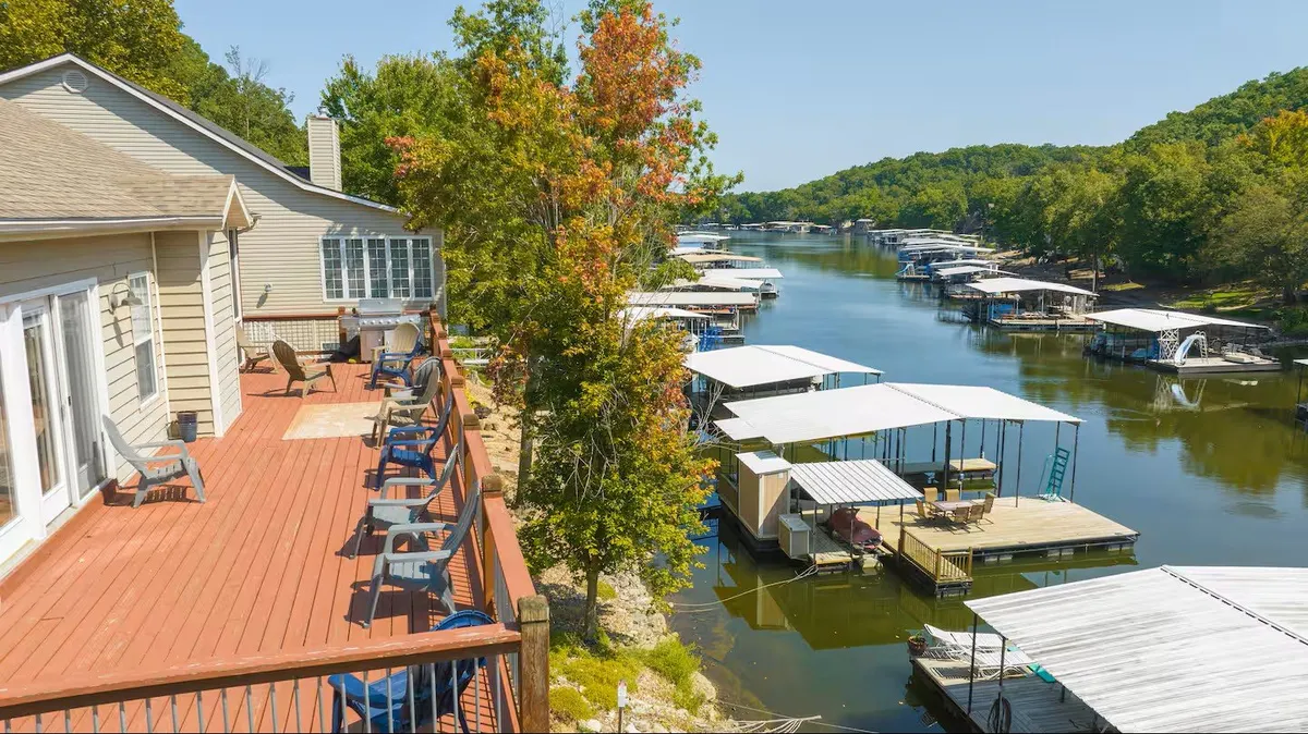 The image showcases the lakehouse's spacious deck furnished with comfortable chairs overlooking a tranquil waterway, framed by lush trees and dotted with boathouses. The scene is set during a serene sunset, suggesting a peaceful, inviting atmosphere perfect for relaxation and gatherings.